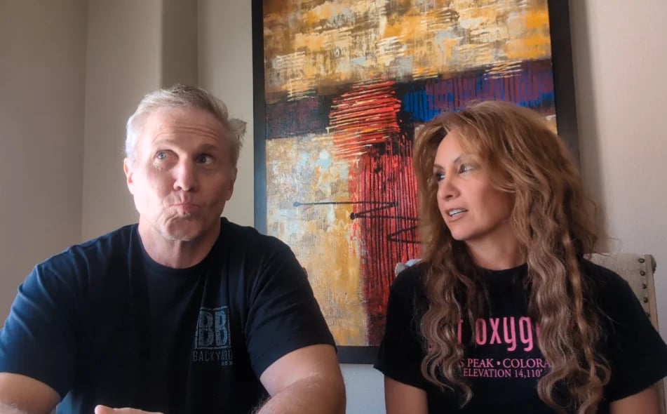 Two people sitting indoors, a man in a dark blue shirt on the left and a woman with wavy hair in a black graphic tee on the right, with abstract artwork hanging on the wall behind them