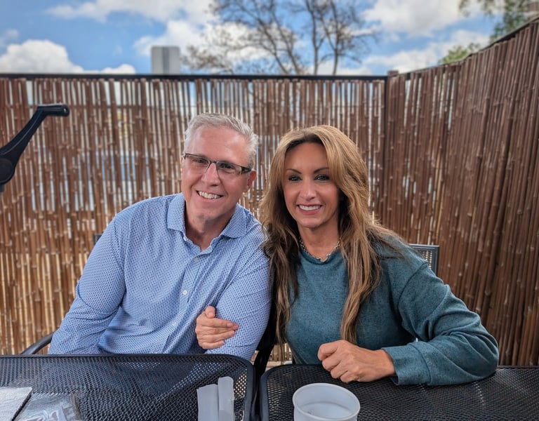 Two smiling adults posing together at an outdoor table with a wooden fence and blue sky in the background